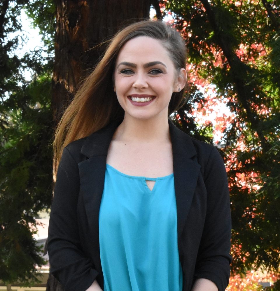 Alicia Martin is wearing a blue shirt and black blazer with a floral and tree filled background.